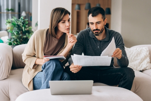 Couple reviewing printed bills and tablet beside laptop on living room sofa