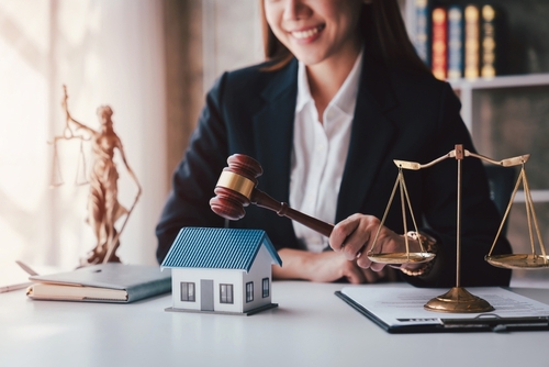 smiling female attorney holding gavel beside scale of justice and house model
